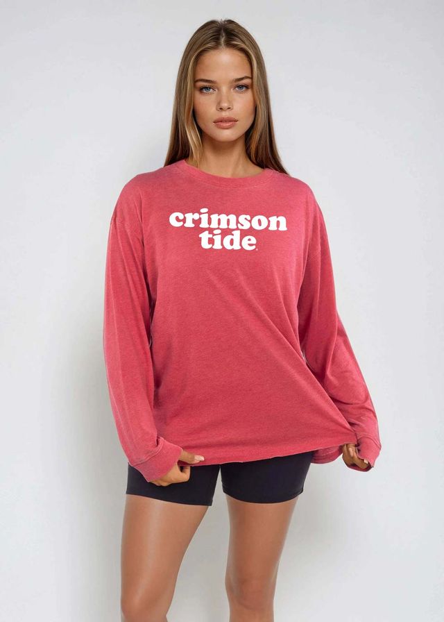 A woman modeling a long-sleeve crewneck shirt in a faded crimson pink with 'crimson tide' printed in white across the chest, paired with black athletic shorts, against a light gray studio background.