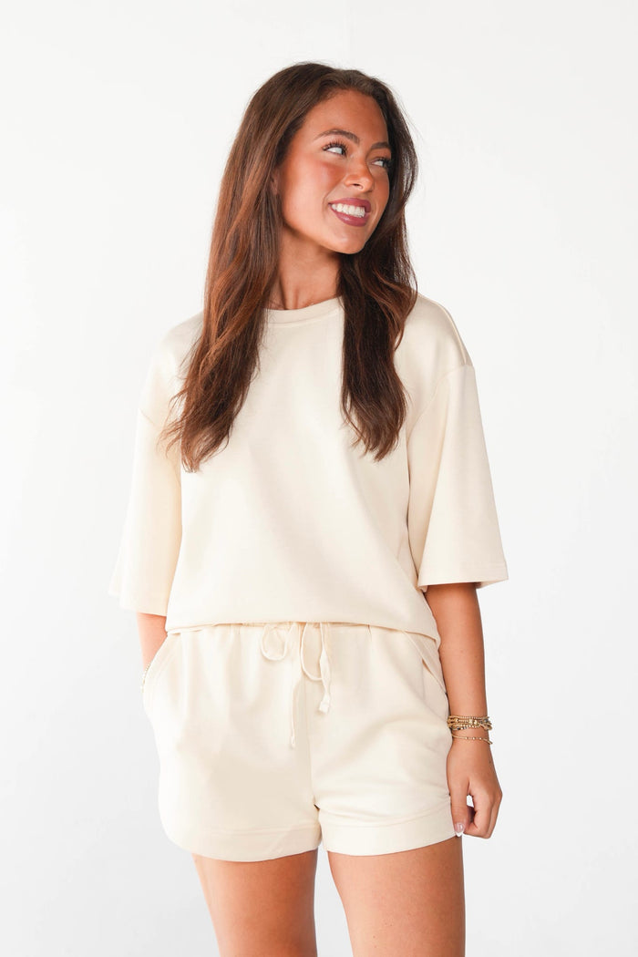 A woman modeling a matching cream-colored lounge set consisting of a short-sleeve crewneck top and drawstring shorts, set against a plain white studio background.