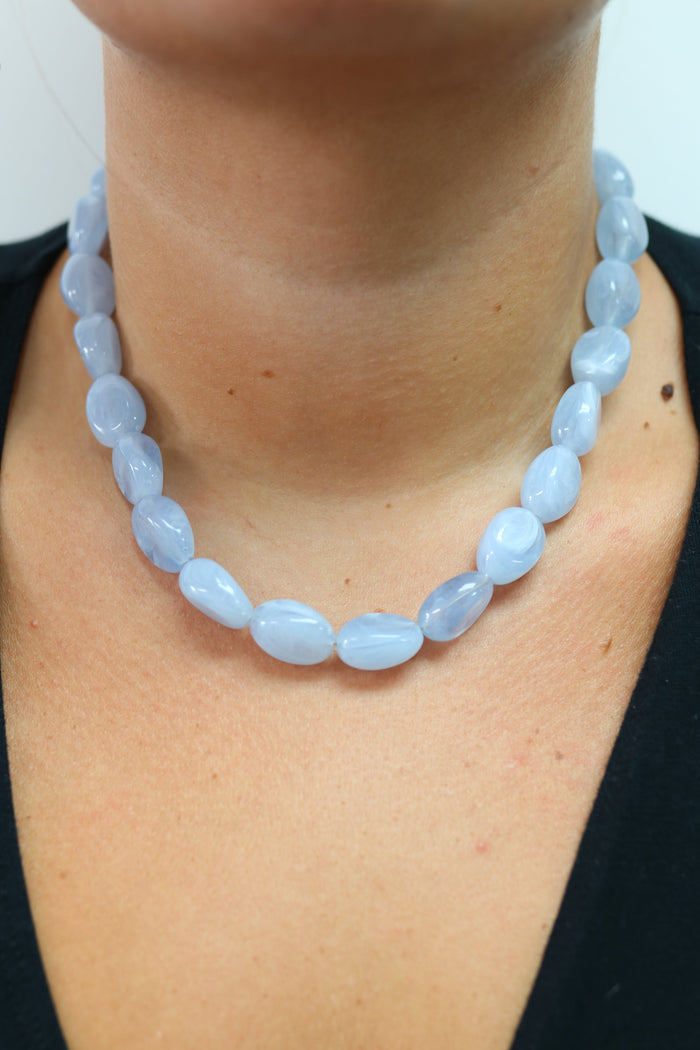 A close-up of a person wearing a light blue beaded necklace with oval-shaped polished beads, paired with a black cardigan and a lavender top.