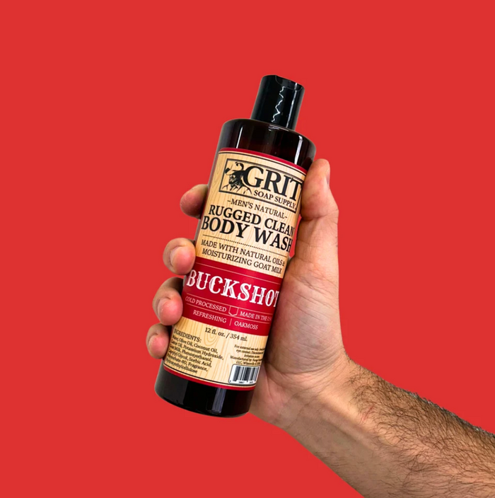 A hand holding a bottle of GRIT Men's Natural Rugged Clean Body Wash against a solid red background.