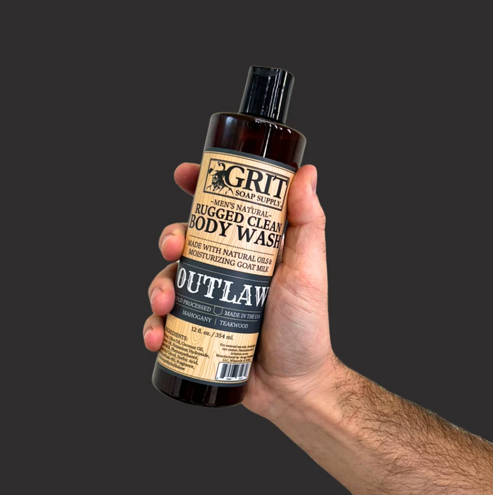 A hand holding a dark amber bottle of Grit Creek Naturals Outlaw Rugged Clean Body Wash against a solid dark gray background.
