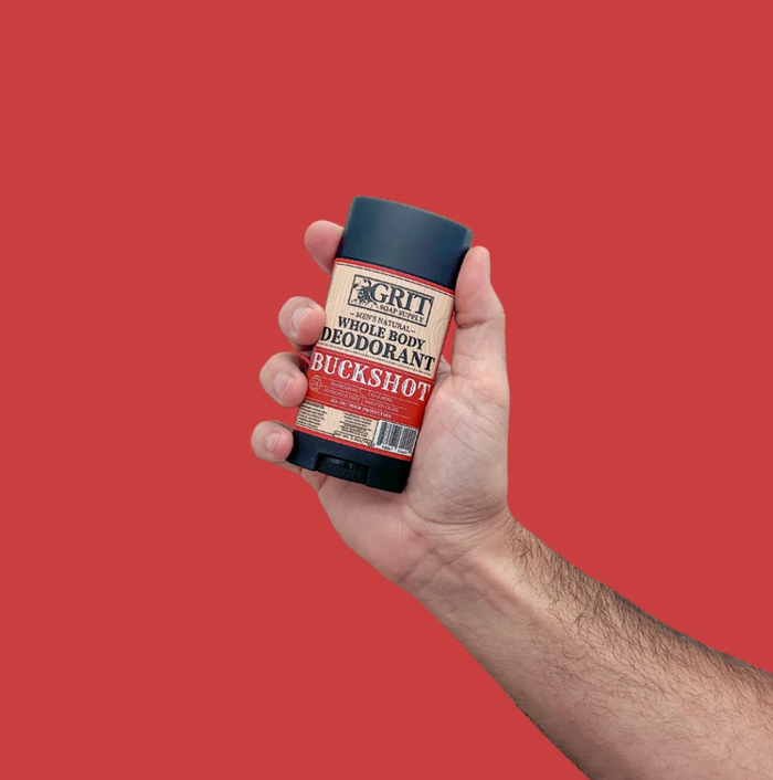 A hand holding a deodorant stick against a solid red background. The product label reads 'CFT Whole Body Deodorant Buckshot'.