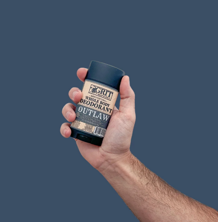 A hand holding a black deodorant stick with a beige and black label that reads 'Grit Outlaw Whole Body Deodorant' against a solid dark slate blue background.