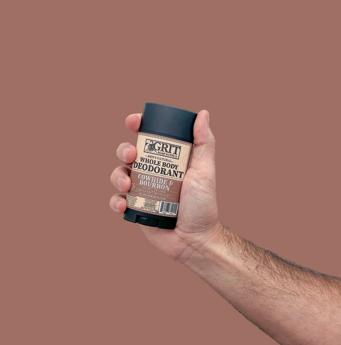 A hand holding a stick of GRIT Whole Body Deodorant in the scent Guilt Free But Robin, against a solid dusty rose background.