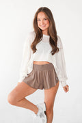 A woman posing in a white cropped long-sleeve shirt and taupe athletic shorts against a white studio background.