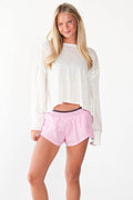 A blonde woman modeling a white long-sleeve cropped sweatshirt and light pink athletic shorts with white sneakers, standing against a plain white studio background.