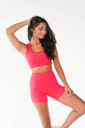 A woman modeling a bright pink activewear set consisting of a scoop-neck crop top and matching high-waisted shorts, posed against a clean white studio background.