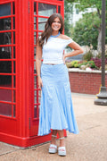 A woman stands outdoors next to a red telephone booth wearing a white off-the-shoulder crop top and a light blue gingham midi skirt, paired with white platform sandals.