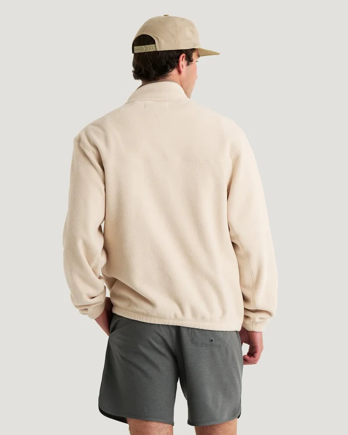 Person wearing a beige jacket and gray shorts with a beige cap on a plain background