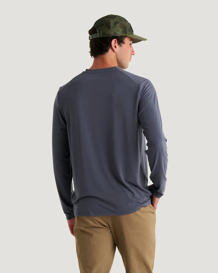 A man shown from behind wearing a slate blue long-sleeve shirt, khaki pants, and a green camouflage baseball cap against a light gray background.