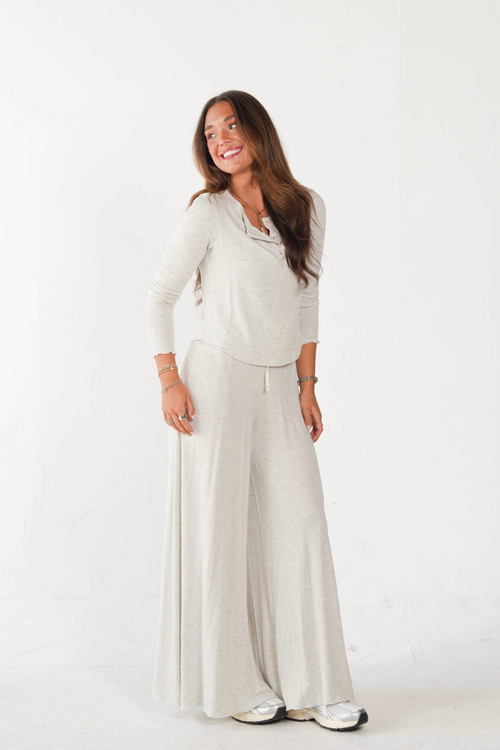 A woman modeling a light beige knit loungewear set consisting of a cowl neck long-sleeve top and matching wide-leg pants, standing against a plain white studio background.