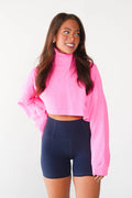 A woman modeling a bright pink long-sleeve crop top with a mock neck and navy blue high-waisted athletic shorts against a plain white background.