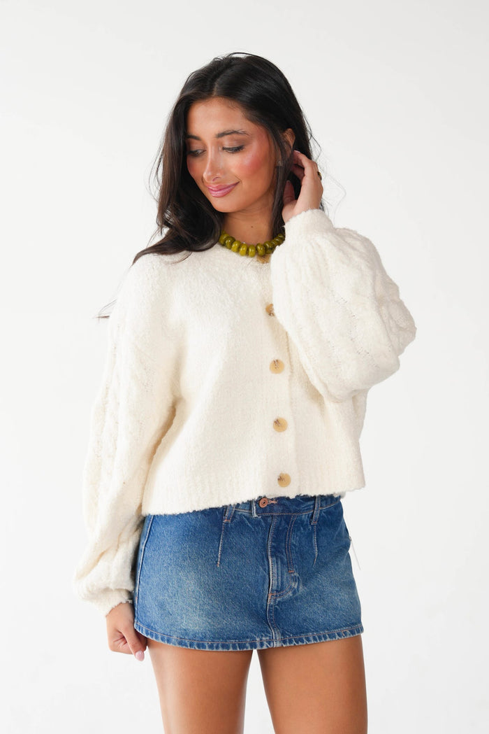 A woman wearing a cream-colored, fuzzy cropped cardigan with wooden buttons and a blue denim mini skirt, standing against a plain white studio background.