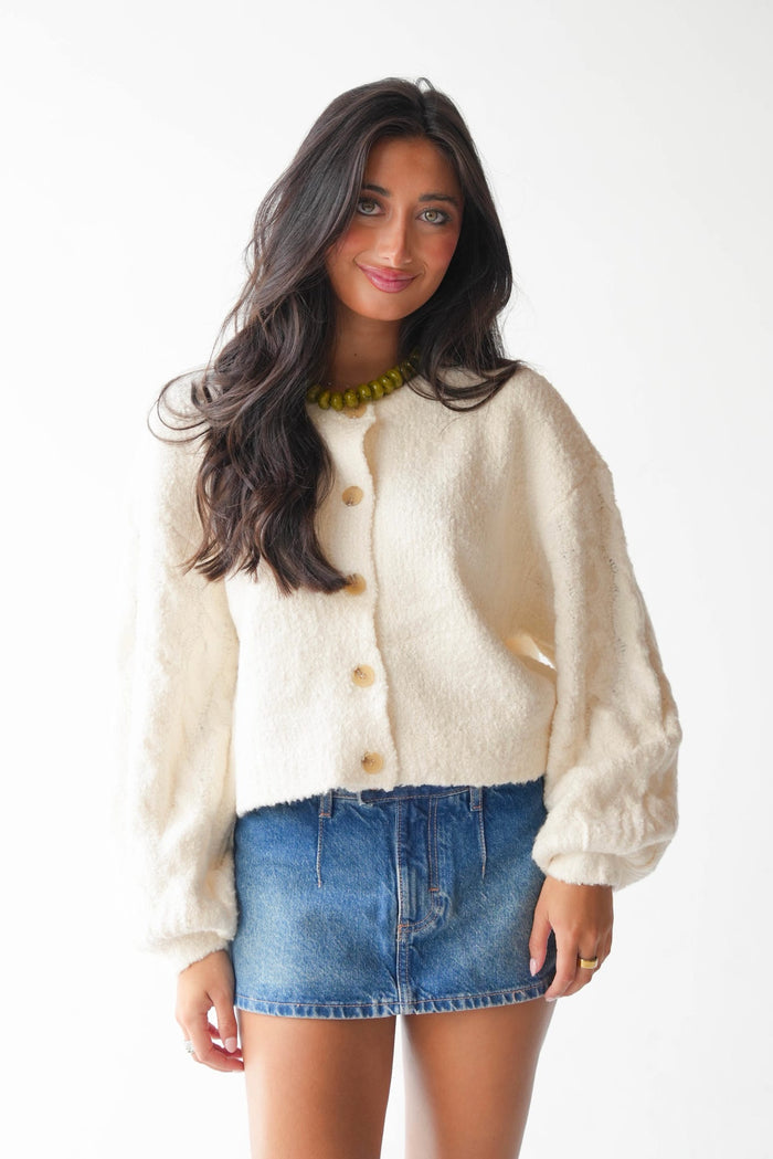 A woman modeling a cream-colored, fuzzy cropped cardigan with gold buttons, paired with a blue denim mini skirt and a green beaded necklace, against a plain white background.