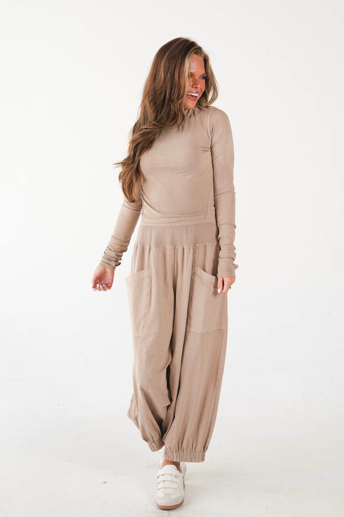 A woman modeling a beige long-sleeve top and matching loose harem-style pants with large front patch pockets, paired with white sneakers, against a plain white studio background.