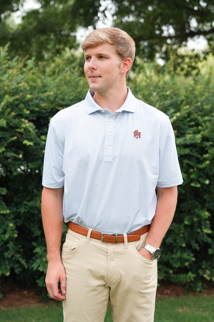 A young man wearing a light blue polo shirt with a small embroidered logo on the left chest, paired with khaki pants and a brown belt, standing outdoors in front of green bushes.