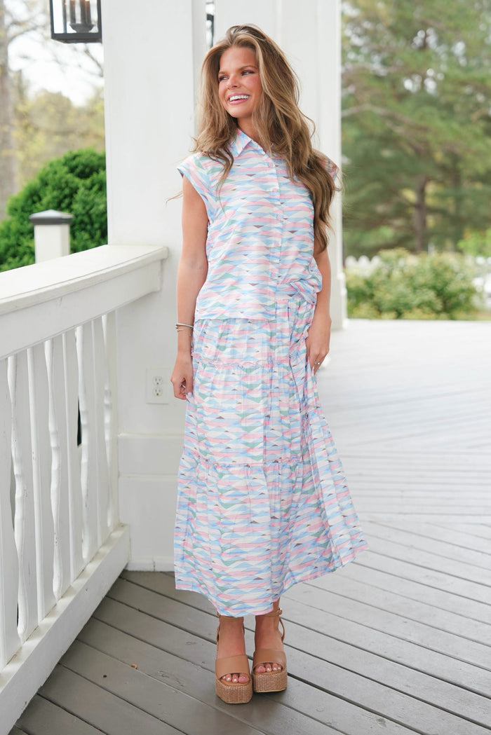 Woman in a striped dress standing on a porch with a white railing and greenery in the background.