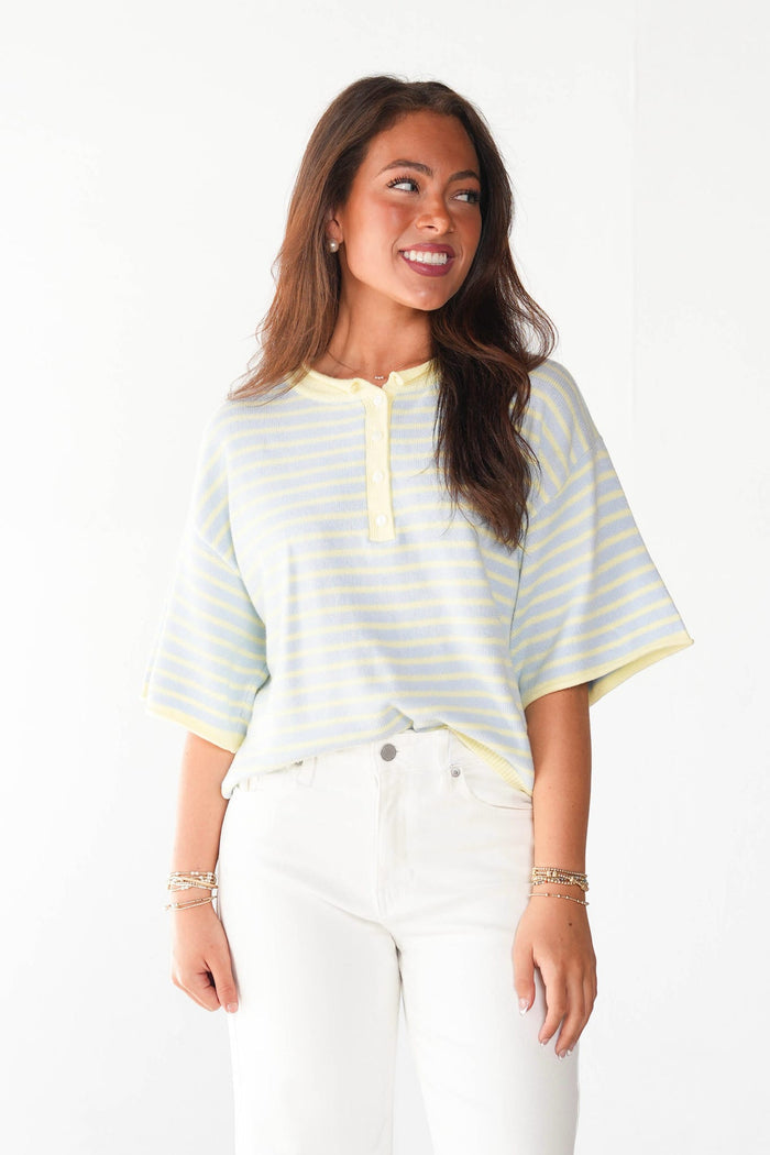 A woman wearing a pale yellow and white striped short-sleeve henley top with white pants, standing against a plain white background.