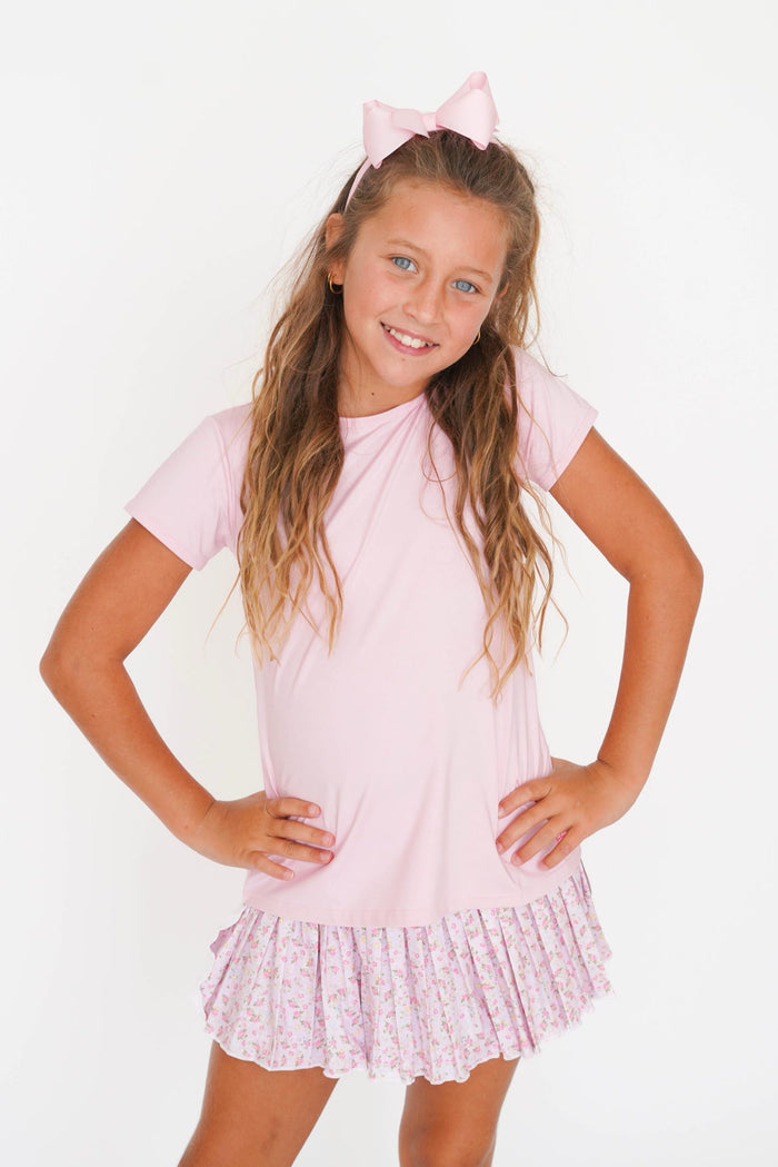 A young girl with long light brown hair wearing a light pink short-sleeve t-shirt dress with a ruffled floral skirt and a matching pink bow headband, posing against a white studio background.