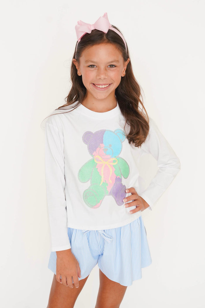 A young girl smiling and posing in a white long-sleeve t-shirt with a colorful pastel teddy bear graphic, paired with light blue and white striped shorts, and a pink bow headband against a plain white background.