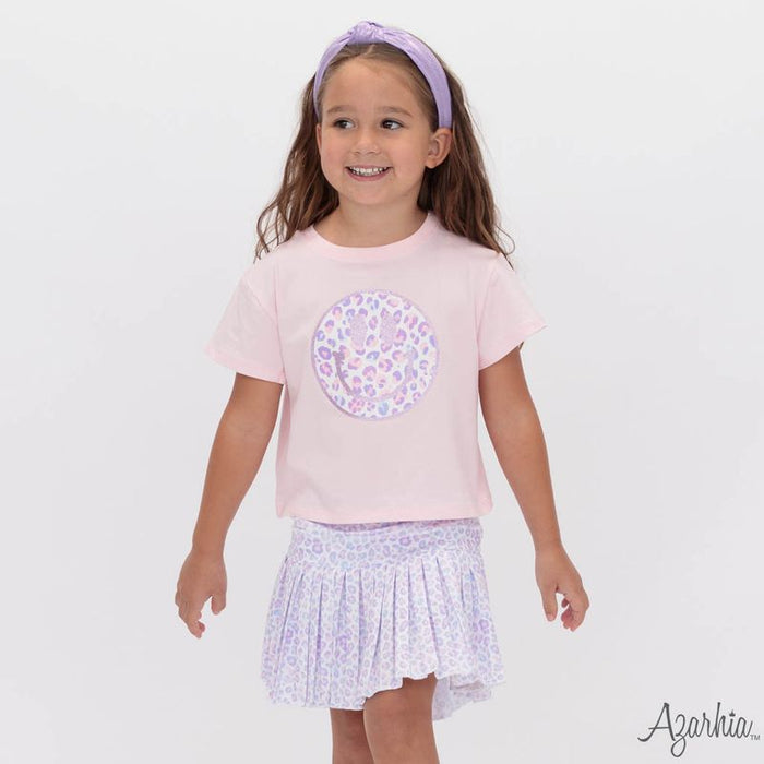A young girl smiling and wearing a light pink short-sleeve t-shirt with a purple and white patterned circular graphic on the chest, paired with a matching light purple patterned ruffled skirt and a lavender headband. The brand name 'Agarkia.