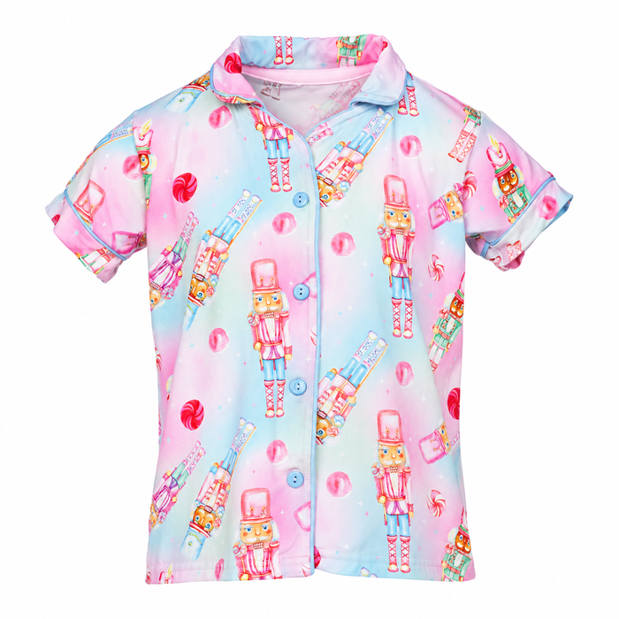 A short-sleeve button-up shirt with a colorful candy and nutcracker print on a pastel pink and blue background, shown against a plain white background.