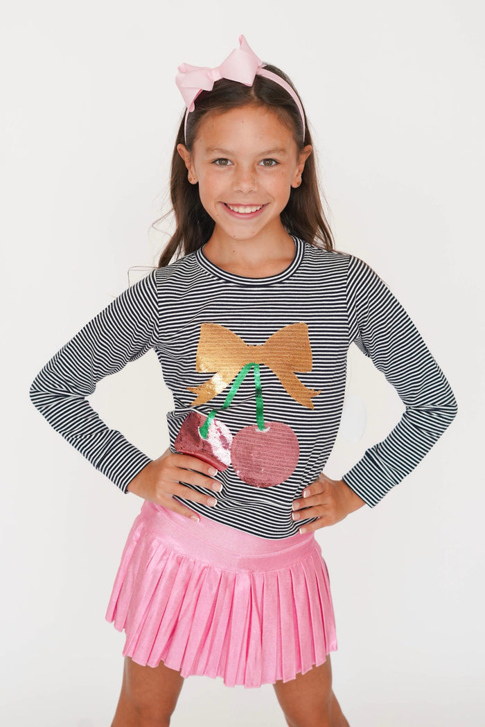 A young girl smiling and posing in a black and white striped long-sleeve top with a sequined cherry and bow graphic, paired with a pink pleated skirt and a pink bow headband, against a plain white background.