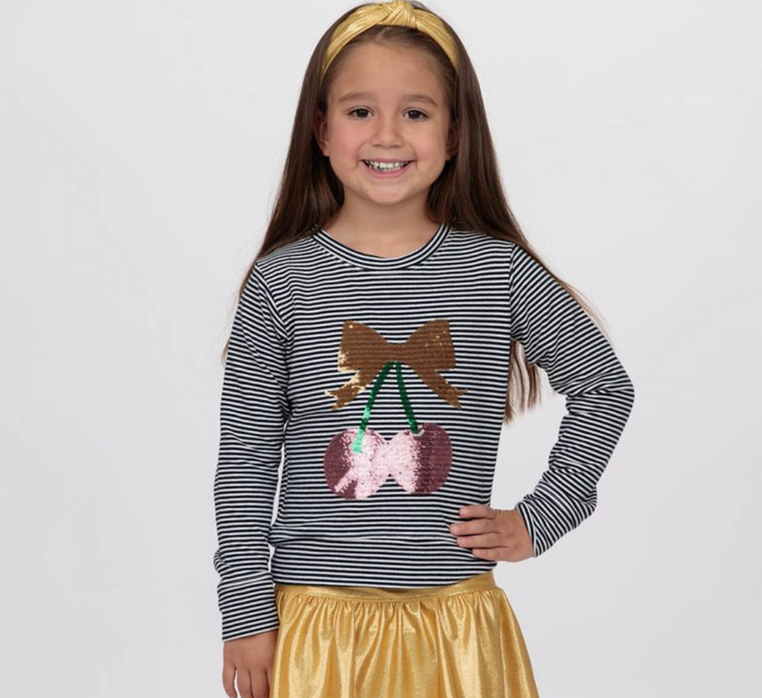 A young girl with a gold headband and a shiny gold skirt is wearing a long-sleeve shirt with black and white horizontal stripes and a sequined cherry graphic on the front.