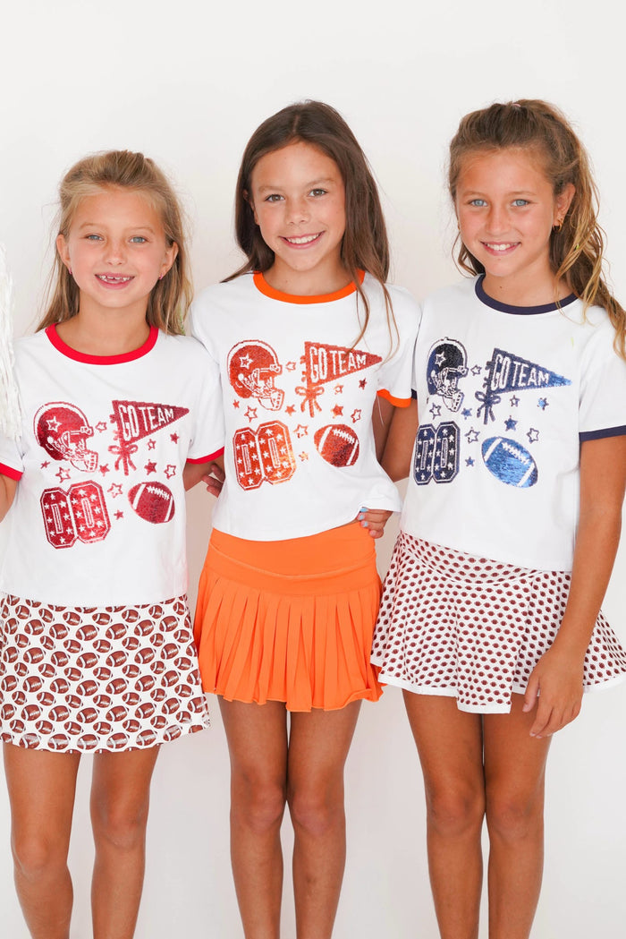 Three young girls standing together and smiling, wearing white short-sleeve athletic-style T-shirts with sports graphics and patterned skirts against a plain white background.