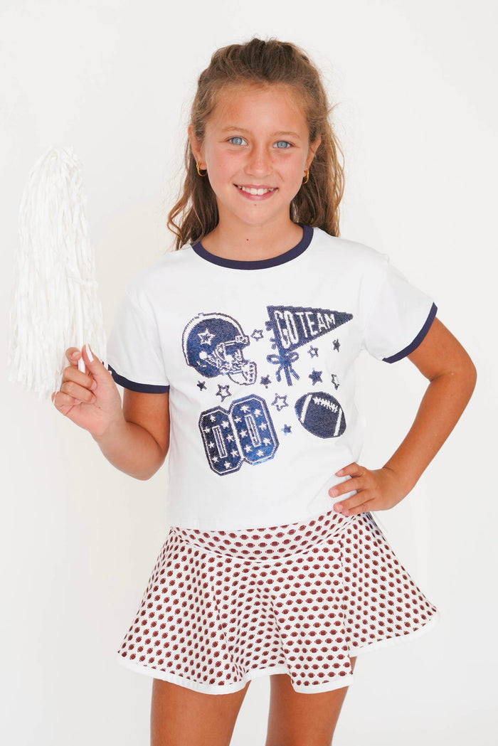 A young girl smiling and holding a white pom-pom, wearing a white short-sleeve ringer T-shirt with navy blue trim and football graphics, paired with a brown and white polka dot skirt, against a plain white background.