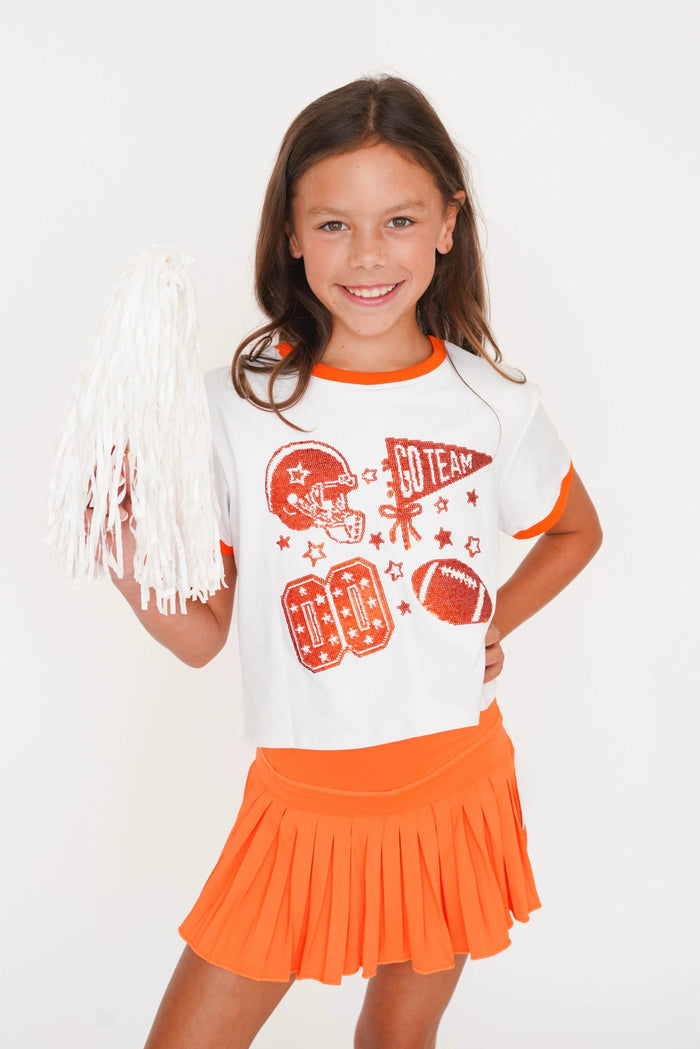 A young girl smiling and holding a white pom-pom, wearing a white ringer T-shirt with orange trim and football graphics, paired with an orange pleated skirt, against a plain white background.