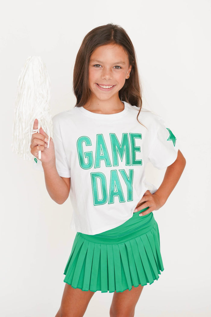 A young girl smiling and holding a white pom-pom, wearing a white t-shirt with 'GAME DAY' printed in green and a green pleated skirt, against a plain white background.