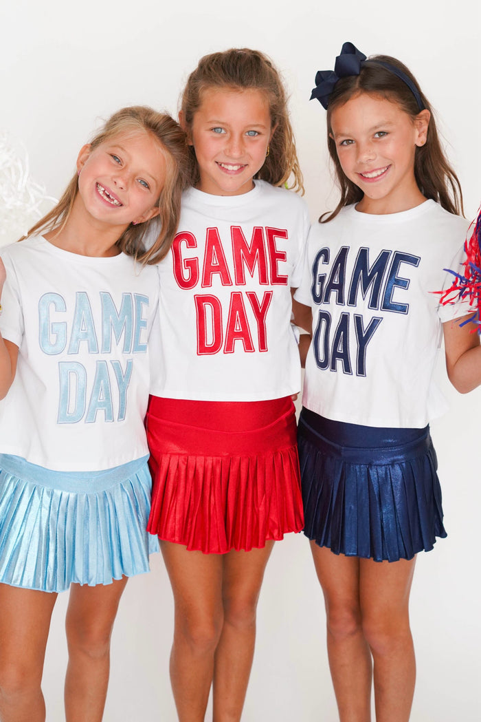 Three young girls wearing matching white 'GAME DAY' graphic t-shirts and pleated skirts in light blue, red, and navy, standing together against a white background.