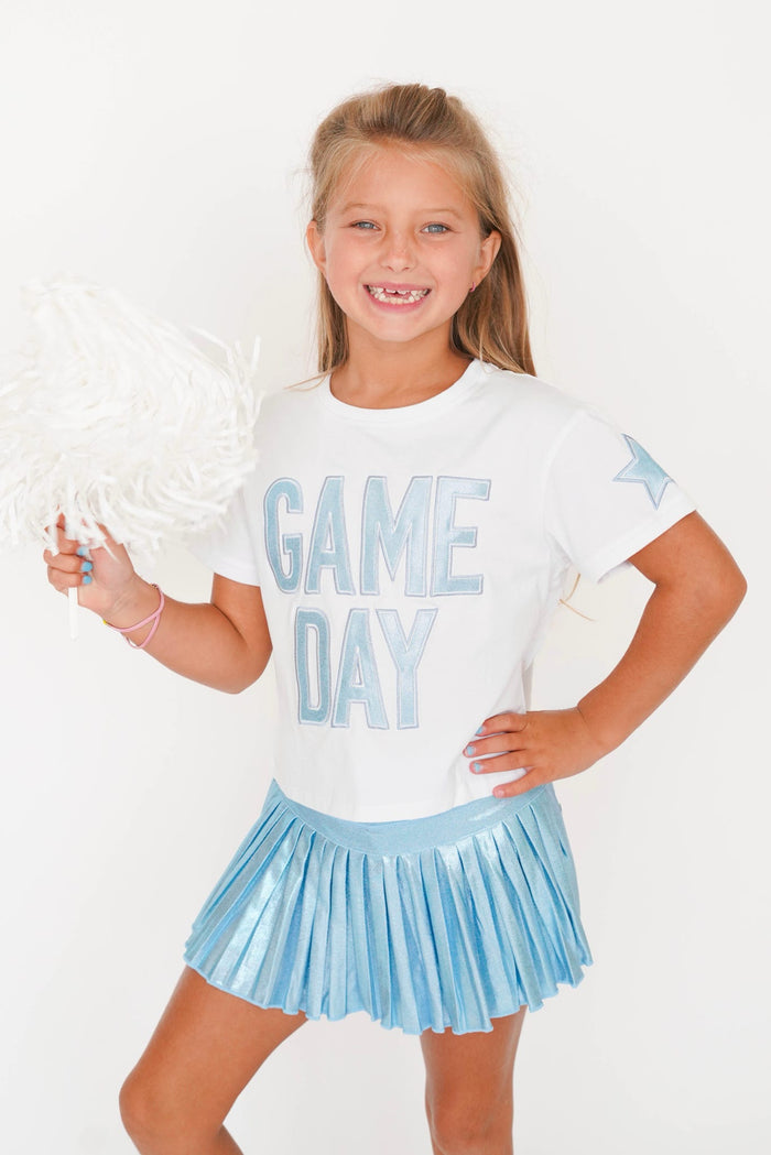 A young girl smiling and posing in a white t-shirt with 'GAME DAY' printed in light blue, paired with a shiny light blue pleated cheerleader skirt. She is holding a white pom-pom against a clean white background.