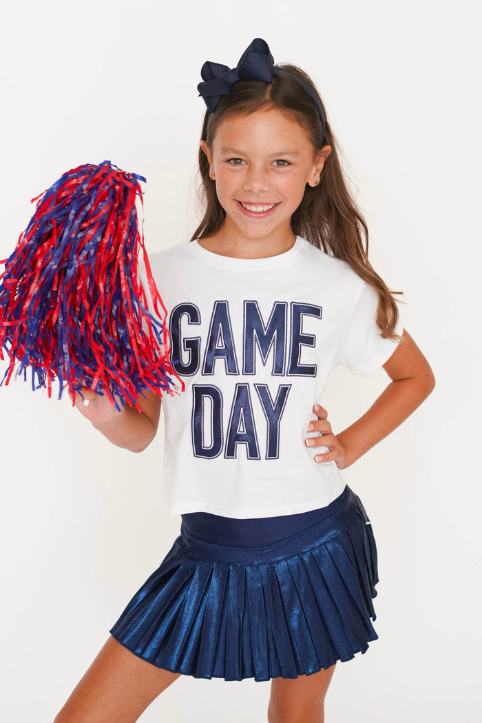 A young girl cheerleader posing in a white t-shirt that says 'GAME DAY' in navy blue, a navy blue pleated skirt, and holding a red and navy blue pom-pom against a plain white background.