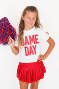 A young girl cheerleader posing in a white t-shirt with 'GAME DAY' printed in red, a red pleated skirt, and holding a red and blue pom-pom against a white background.