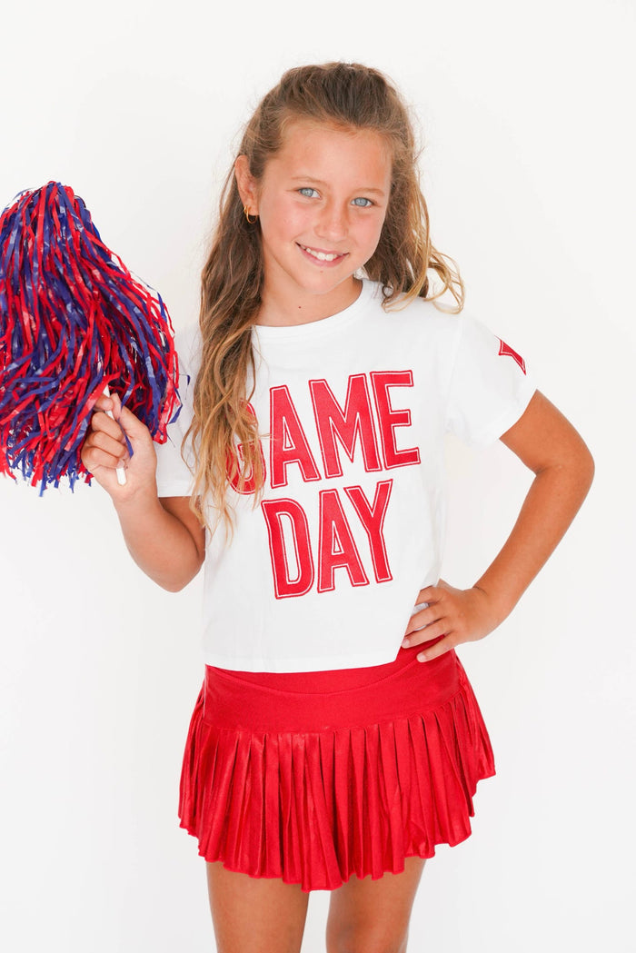 A young girl cheerleader posing in a white t-shirt with 'GAME DAY' printed in red, a red pleated skirt, and holding a red and blue pom-pom against a white background.