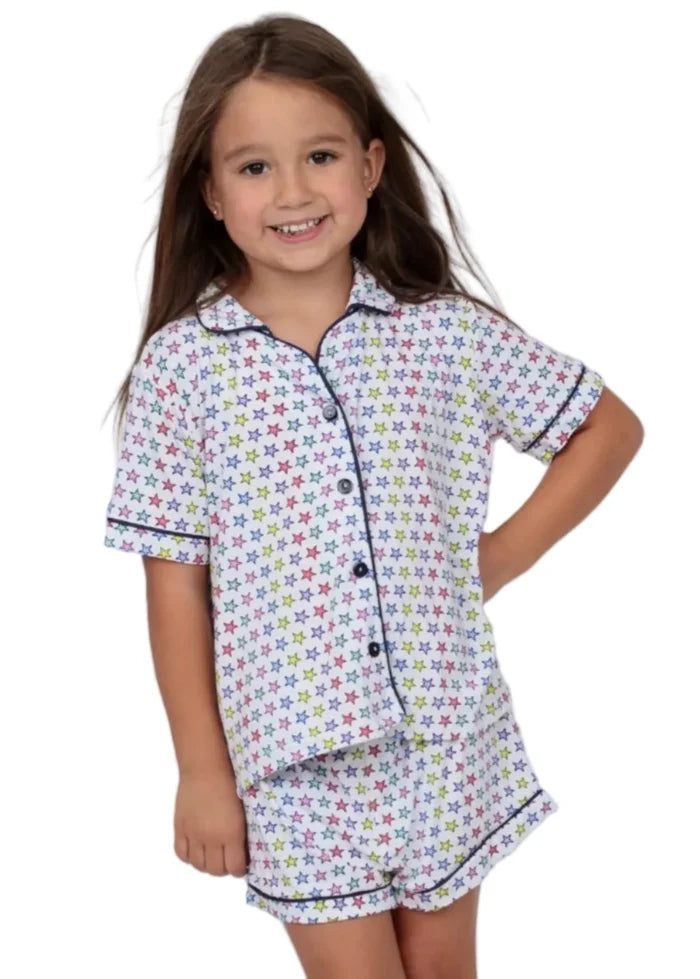 A young girl with long brown hair smiling while wearing a two-piece pajama set with a colorful star pattern on a white background, set against a solid light blue backdrop.