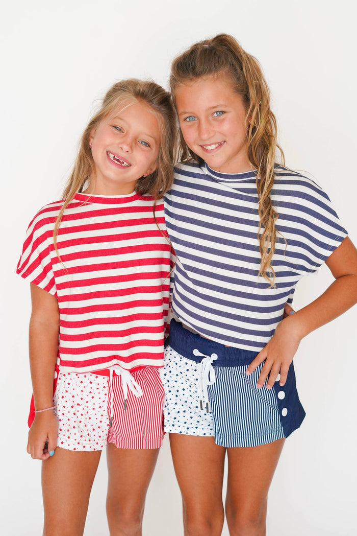Two young girls smiling and posing together in matching nautical-themed striped t-shirts and shorts. The girl on the left wears a red and white striped shirt with pink polka-dot shorts, and the girl on the right wears a navy and white striped shirt with navy and white striped shorts.
