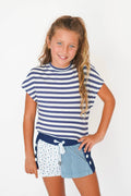 A young girl with blonde hair in a ponytail, smiling and posing in a navy and white striped t-shirt and matching patterned shorts against a plain white background.