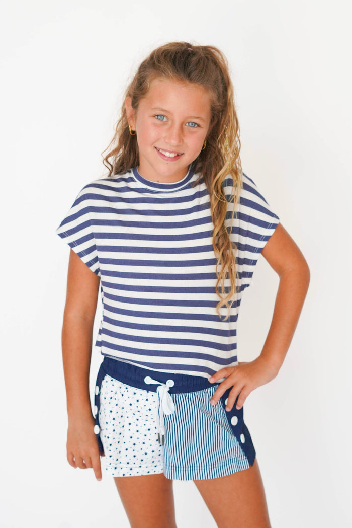 A young girl with blonde hair in a ponytail, smiling and posing in a navy and white striped t-shirt and matching patterned shorts against a plain white background.
