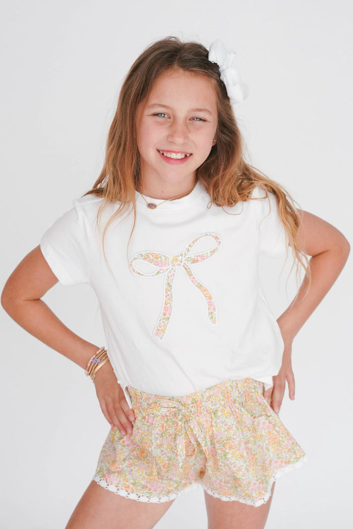 Young girl wearing a white top with a bow and floral shorts on a plain background