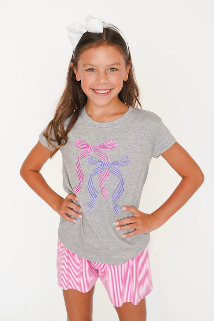 A young girl with a white bow in her hair models a heather gray t-shirt featuring a pink and purple ribbon ballet dancer graphic, paired with pink and white striped shorts against a plain white background.