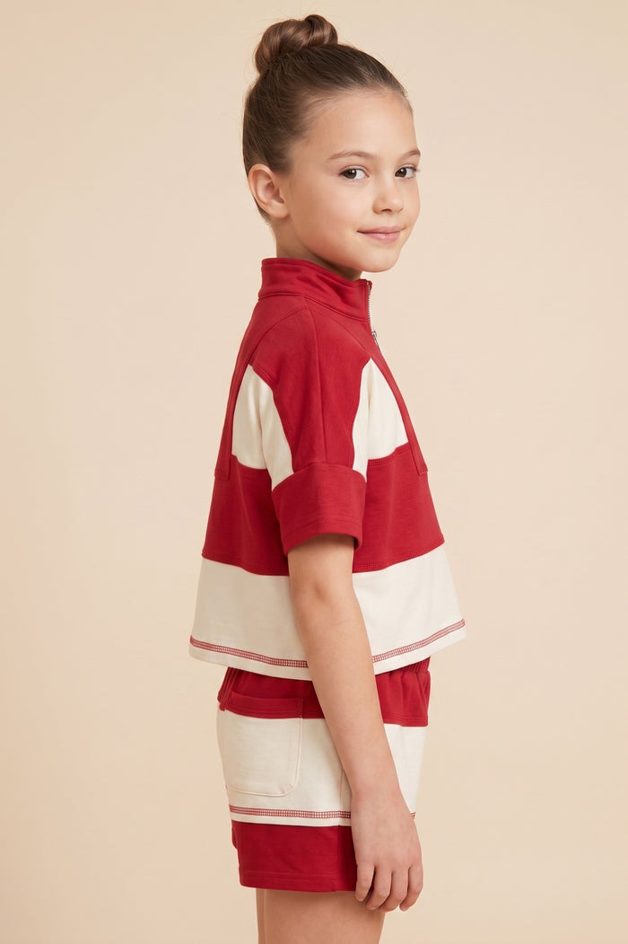 A young girl with her hair in a bun is shown from the side, wearing a red and off-white striped short-sleeve mock-neck top and matching shorts against a solid beige background.