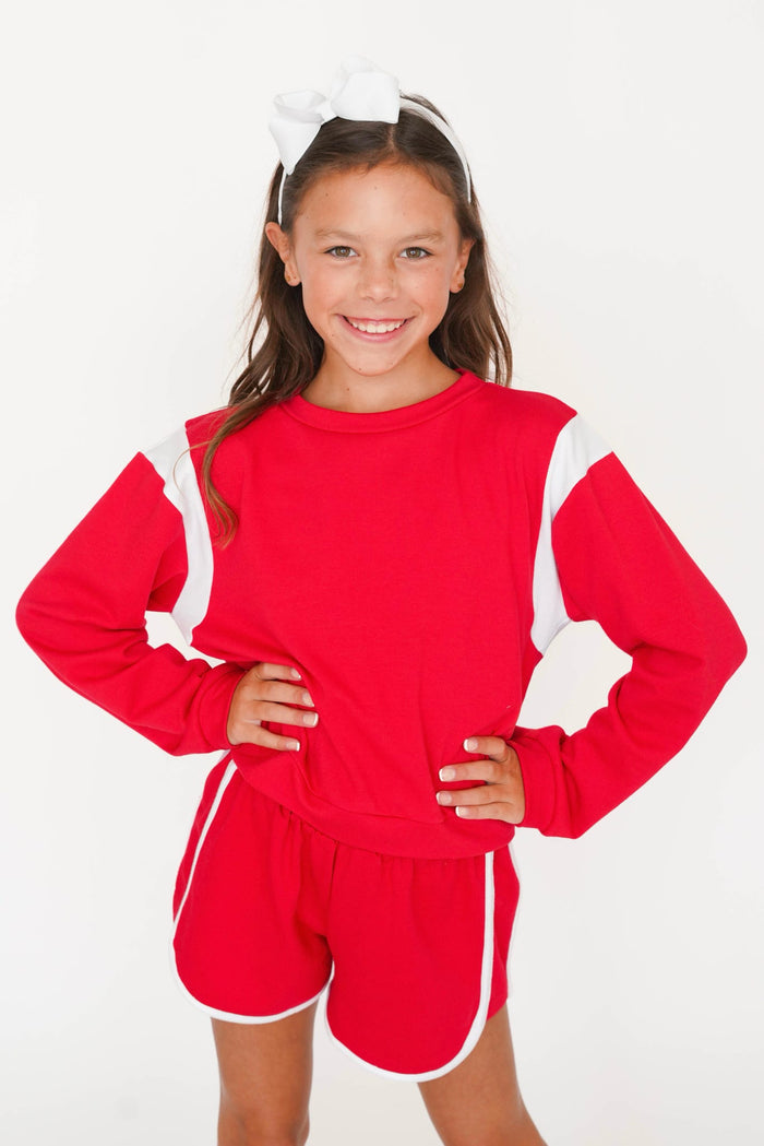 A young girl smiling and posing in a matching red and white athletic lounge set, featuring a long-sleeve sweatshirt and shorts, with a white bow headband against a plain white background.