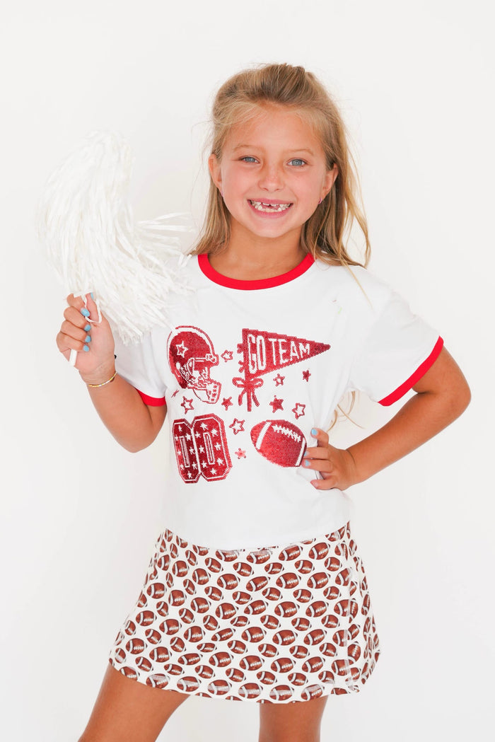 A young girl smiling and posing in a white and red athletic-themed t-shirt and a matching white skirt with football patterns. She is holding a white pom-pom against a clean white background.
