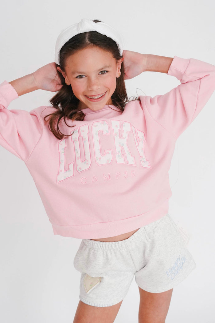 Young girl wearing a pink sweatshirt with 'LUCKY' text and white shorts on a light gray background