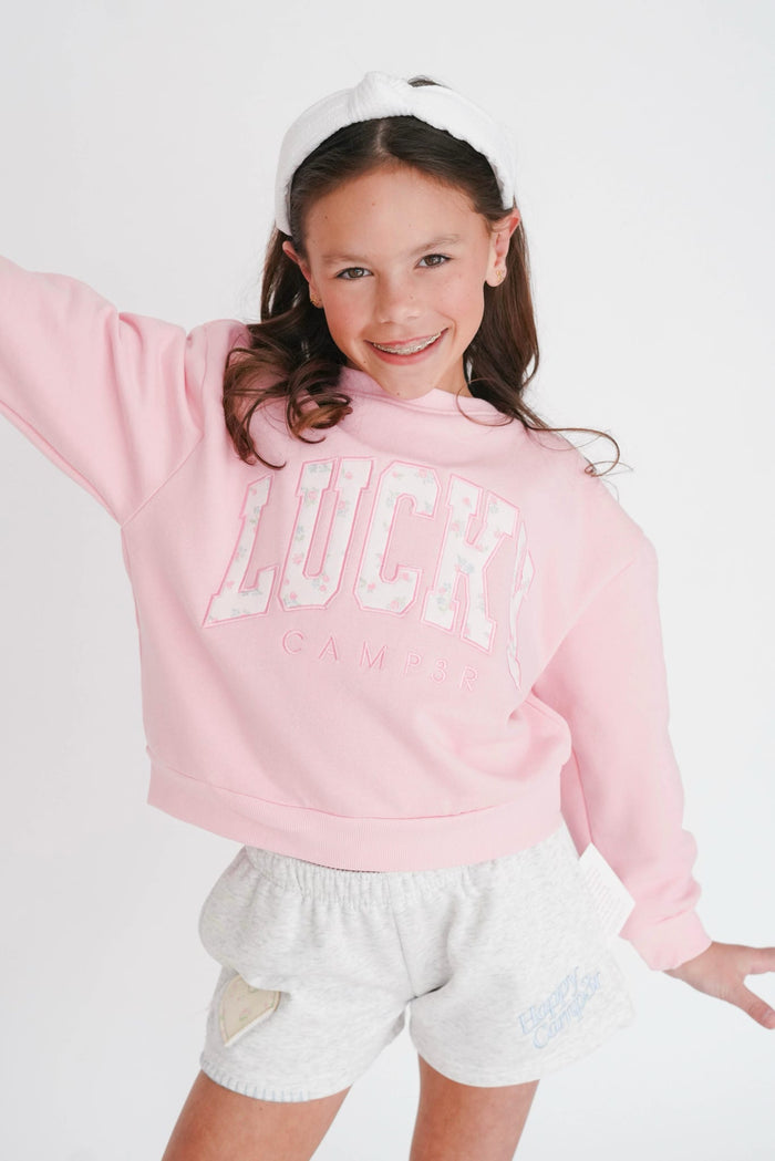 Young girl wearing a pink sweatshirt with 'LUCK' and white shorts on a white background
