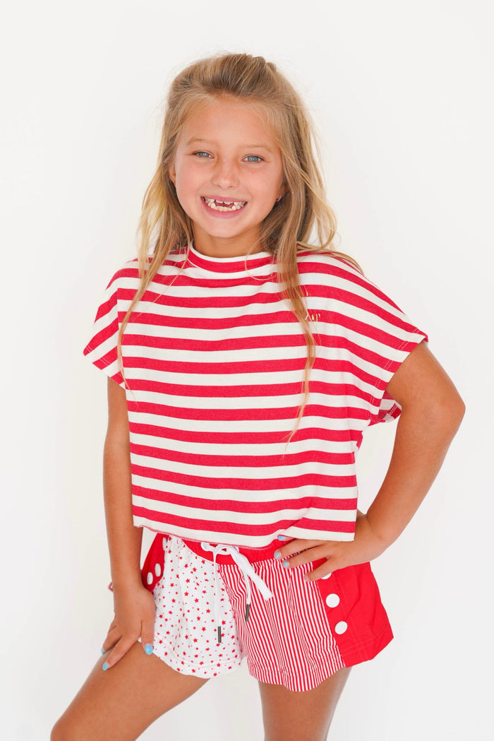 A young girl smiling and posing in a red and white striped t-shirt and matching red, white, and polka dot shorts against a plain white background.