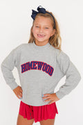 A young girl smiling and posing in a heather gray crewneck sweatshirt with 'HOMEWOOD' printed in red and blue arched lettering across the chest, paired with a red pleated skirt and a navy blue hair bow.